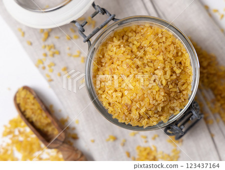 Glass jar of raw dry uncooked yellow bulgur grain with a wooden scoop on napkin top view 123437164