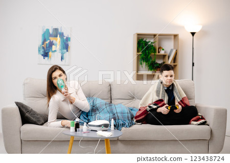 Young woman holding a mask nebulizer and inhaling fumes medication into lungs. Sick lady inhaling through inhaler mask at home and funny boy with gamepad playing video game at home near her Young woman holding a mask nebulizer and inhaling fumes medication into lungs. Sick lady inhaling through inhaler mask at home and funny boy with gamepad playing video game at home near her 123438724