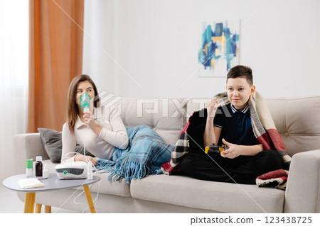 Young woman holding a mask nebulizer and inhaling fumes medication into lungs. Sick lady inhaling through inhaler mask at home and funny boy with gamepad playing video game at home near her Young woman holding a mask nebulizer and inhaling fumes medication into lungs. Sick lady inhaling through inhaler mask at home and funny boy with gamepad playing video game at home near her 123438725