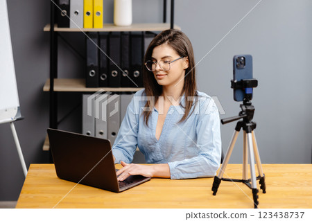 Videoconference And Training. Class of diverse students learning remotely online using web app. Female teacher providing lesson with laptop, recording video on cellphone on tripod 123438737