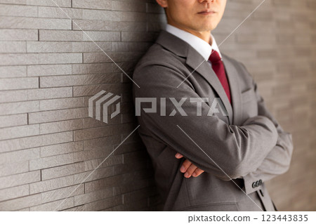 A middle-aged male businessman in a gray suit leaning against a wall with his arms folded A middle-aged male businessman in a gray suit leaning against a wall with his arms folded 123443835
