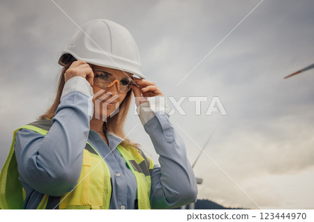 A professional female engineer is meticulously adjusting her safety gear while working on a large wind farm, ensuring both her safety and efficiency in her role within the renewable energy sector 123444970