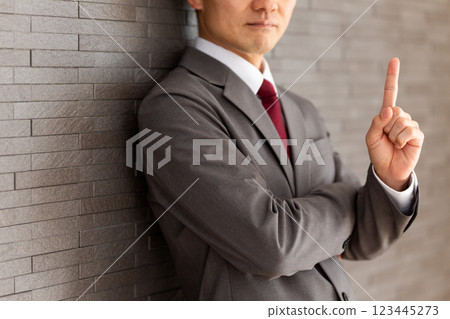 一位身穿灰色西裝的中年男性商人靠在牆上，用食指指著 123445273