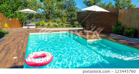 Modern backyard pool with turquoise water, wooden deck, umbrellas, lounge chairs, and pink inflatable ring 123445760
