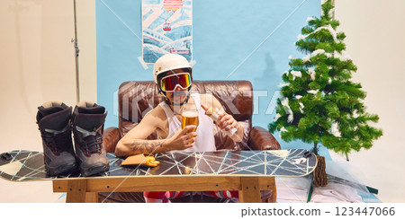 Man in ski goggles enjoying beer and hot dog, sitting on leather couch at home with Christmas tree in background. 123447066