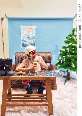 Quirky man in helmet and goggles enjoying beer and hot dog, sitting at wooden table with snowboard against studio background. 123447067