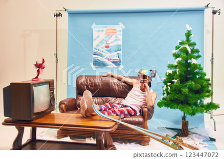 Man reclining on brown leather couch in plaid pajama pants, ski resting on wooden table, wearing ski goggles with humorous beverage helmet 123447072