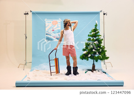 Man wearing white tank top, underwear, fur hat and boots standing with sleds against abstract winter background with artificial snow and tree. 123447096