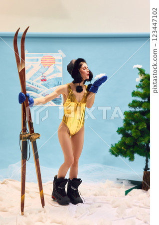 Full-length portrait of happy young woman in golden swimsuit and fur hat, standing in boots, holding skis and blowing snow from hand 123447102
