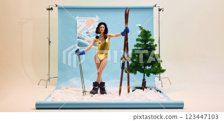 Young woman in gold swimsuit and ski boots confidently standing against winter-themed backdrop. 123447103