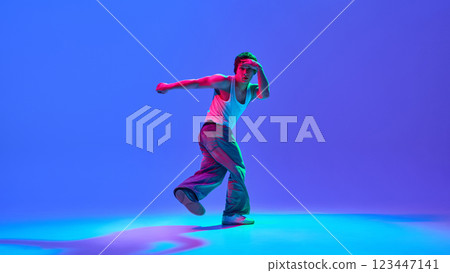Dynamic image of male performance, breakdance in casual attire in motion against gradient blue purple background in neon light Dynamic image of male performance, breakdance in casual attire in motion against gradient blue purple background in neon light 123447141