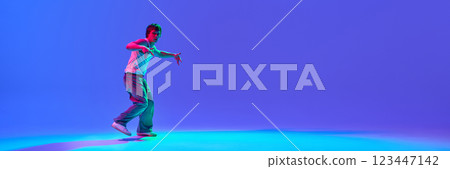 Dynamic image of young guy in casual clothes dancing hip-hop against gradient blue purple background in neon light 123447142