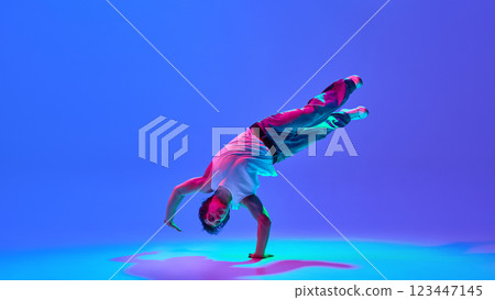 Dynamic young performer executing powerful breakdance move, blending skill and rhythm against neon blue purple backdrop. 123447145