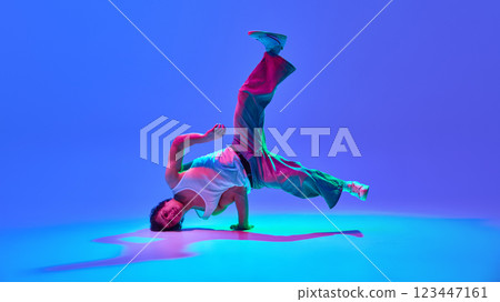 Talented and energetic young man, dancer in motion, performing breakdance against gradient blue purple background in neon light 123447161