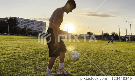 Young man juggling soccer ball on stadium at sunset. Professional footballer kicking ball at green field. Sportsman practicing tricks at meadow with sunlight at background. Freestyle football 123448349