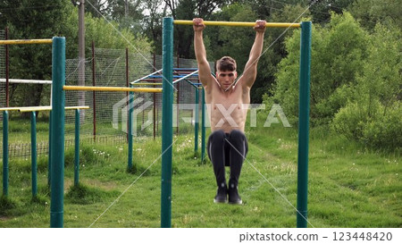 Strong guy training abdominal muscles on horizontal bar at sports ground. Young sportsman exercising on pull-up bar outdoor. Sporty athlete working out at nature. Concept of sportive lifestyle Strong guy training abdominal muscles on horizontal bar at sports ground. Young sportsman exercising on pull-up bar outdoor. Sporty athlete working out at nature. Concept of sportive lifestyle 123448420