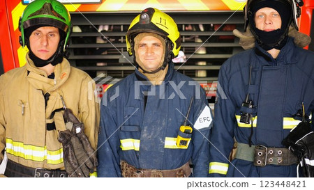 Portrait of young happy fireguards in protective uniforms against background of fire truck at station. Male firefighters in full equipment looking into camera with positive emotions near big red car Portrait of young happy fireguards in protective uniforms against background of fire truck at station. Male firefighters in full equipment looking into camera with positive emotions near big red car 123448421