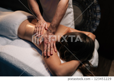 Professional masseur performing relaxing back massage on female client lying on massage table, focusing on shoulder blades and applying oil in spa center with dark interior. Concept of luxury massage. 123451229