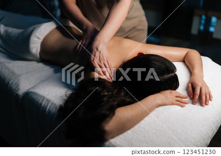 Cropped shot of skilled massage therapist working on female client shoulders muscles during therapeutic session, providing deep tissue relaxation in serene, softly illuminated wellness environment. 123451230