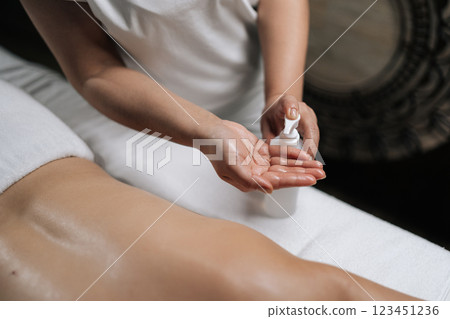 High-angle view of professional masseuse pouring massage oil from white dispenser onto hands, preparing for relaxing body treatment in wellness center with dark interior. Concept of luxury massage. 123451236
