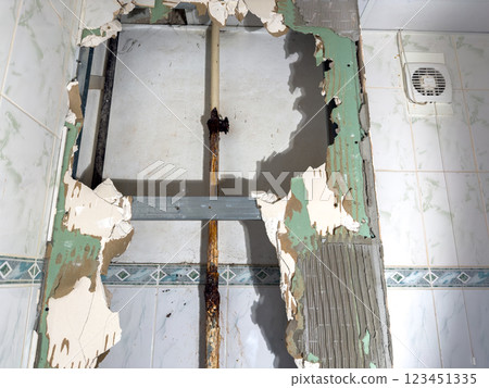 A detailed closeup view of a wall that has been significantly damaged, inside it a visible pipe with rust at plumbing joint and broken tiles 123451335