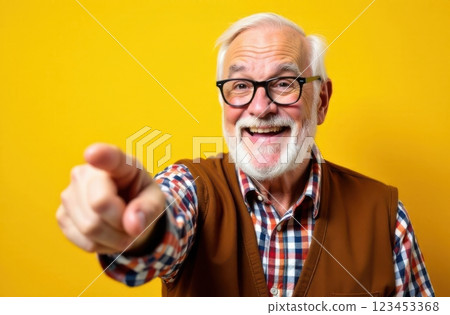 A cheerful senior man with glasses and a white beard stands against a vibrant yellow backdrop. He wears a plaid shirt with a vest and shares a warm joyful expression while pointing. 123453368