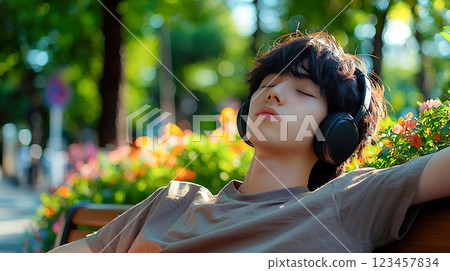 年輕人坐在明亮的公園長椅上，戴著耳機閉著眼睛聽音樂 123457834