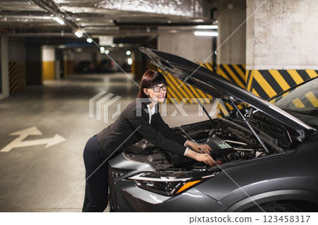 Caucasian businesswoman in formal attire fixes car engine in dimly lit parking garage, conveys independence and problem-solving skills. Caucasian businesswoman in formal attire fixes car engine in dimly lit parking garage, conveys independence and problem-solving skills. 123458317