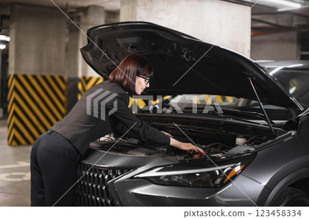 Caucasian businesswoman repairs car engine in underground parking lot, showcasing independence and empowerment. Image captures modern woman in professional attire solving mechanical issue. Caucasian businesswoman repairs car engine in underground parking lot, showcasing independence and empowerment. Image captures modern woman in professional attire solving mechanical issue. 123458334