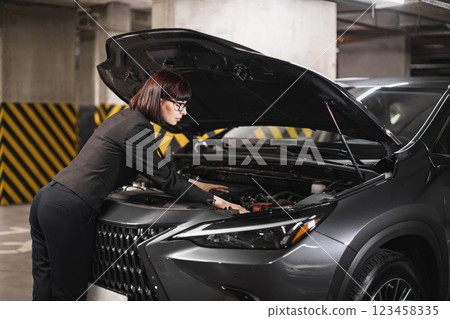 Caucasian businesswoman repairs car engine in underground parking lot, showcasing independence and empowerment. Image captures modern woman in professional attire solving mechanical issue. Caucasian businesswoman repairs car engine in underground parking lot, showcasing independence and empowerment. Image captures modern woman in professional attire solving mechanical issue. 123458335