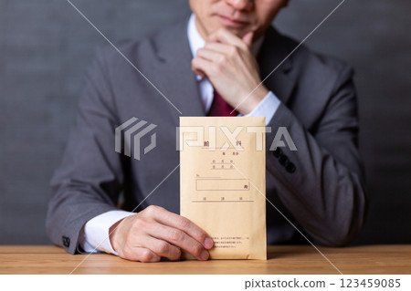 A middle-aged male businessman wearing a gray suit and holding a pay envelope is worried 123459085