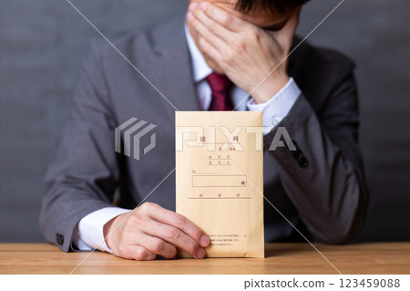 A middle-aged male businessman wearing a gray suit and holding a pay envelope is worried 123459088