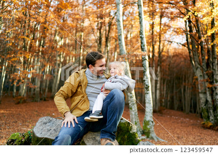Smiling dad with a little girl on his knees sits on a stump Smiling dad with a little girl on his knees sits on a stump 123459547