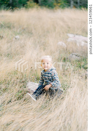 Little smiling girl sits on a large flat stone in tall dry grass Little smiling girl sits on a large flat stone in tall dry grass 123459661
