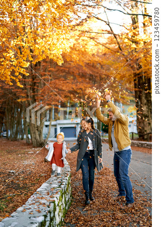 Dad sprinkles dry leaves on mom and little girl walking through the autumn forest 123459780