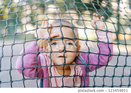 Little girl rests her hands on the tennis net and looks out through its cells Little girl rests her hands on the tennis net and looks out through its cells 123459851