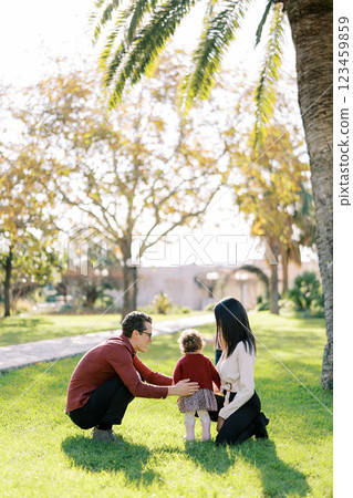 Dad hugs a little girl squatting next to mom in the park. Back view 123459859