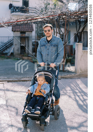 Dad with a little girl in a stroller walks along the road near the house 123460086