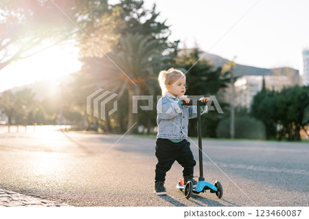 Little girl stands holding the steering wheel of a scooter on the road and looks ahead 123460087