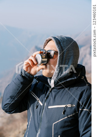 Man in a hooded jacket drinks coffee from a mug of a thermos in the mountains Man in a hooded jacket drinks coffee from a mug of a thermos in the mountains 123460101