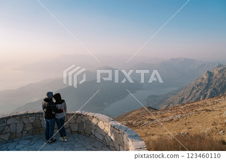 Couple of travelers in jackets hug on an observation deck over the Bay of Kotor in the fog. Montenegro. Back view Couple of travelers in jackets hug on an observation deck over the Bay of Kotor in the fog. Montenegro. Back view 123460110