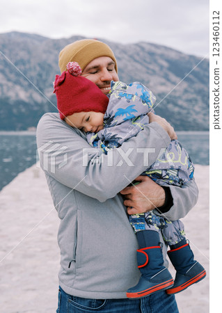 Smiling dad cuddles a little girl on a pier against the backdrop of mountains Smiling dad cuddles a little girl on a pier against the backdrop of mountains 123460112