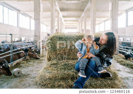 Smiling mom with little girl on her lap sitting on haystack near goat pens at farm Smiling mom with little girl on her lap sitting on haystack near goat pens at farm 123460116