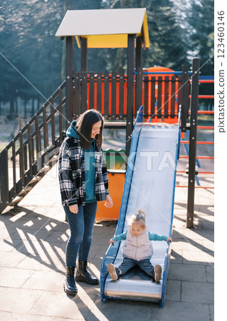 Mom stands next to a little girl sitting on a slide in the playground 123460146
