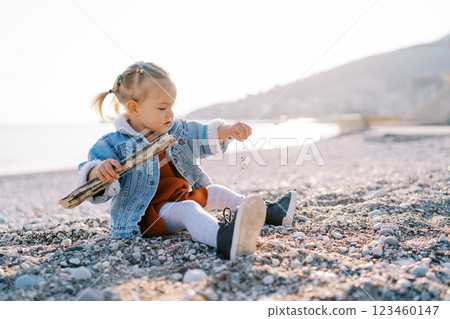 Little girl sits on the beach with a branch and sprinkles small pebbles. Side view Little girl sits on the beach with a branch and sprinkles small pebbles. Side view 123460147