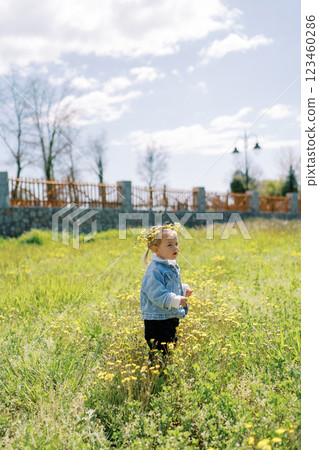 Little girl in a dandelion wreath stands on a flowering lawn. Side view Little girl in a dandelion wreath stands on a flowering lawn. Side view 123460286