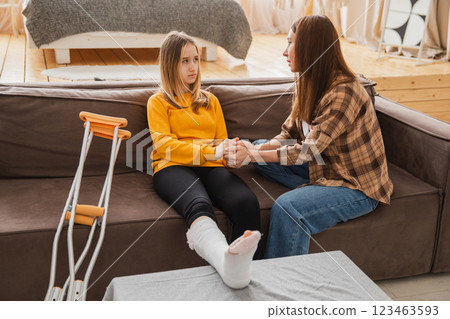 Girl with broken leg in cast sitting with mother on couch at home. Sad teenage kid recovering from broken leg. Girl using cast crutches for quick rehabilitation. Mom supporting daughter Girl with broken leg in cast sitting with mother on couch at home. Sad teenage kid recovering from broken leg. Girl using cast crutches for quick rehabilitation. Mom supporting daughter 123463593