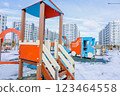 A bright children's playground for kids with slides, cars and stairs stands in the snow against the background of a residential complex of multi-storey modern houses of white and gray color with large 123464558