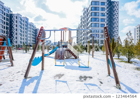A children's playground with swings, slides and stairs stands in the snow against the background of a residential complex of multi-storey modern houses of white and gray color with large windows. High 123464564