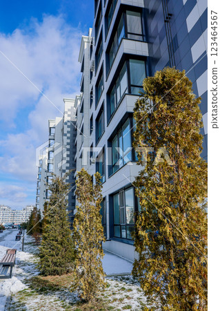 Side view of a multi-storey residential building of modern architecture with large windows against the background of the sky with clouds, the house is lined with white and gray slabs, coniferous Side view of a multi-storey residential building of modern architecture with large windows against the background of the sky with clouds, the house is lined with white and gray slabs, coniferous 123464567